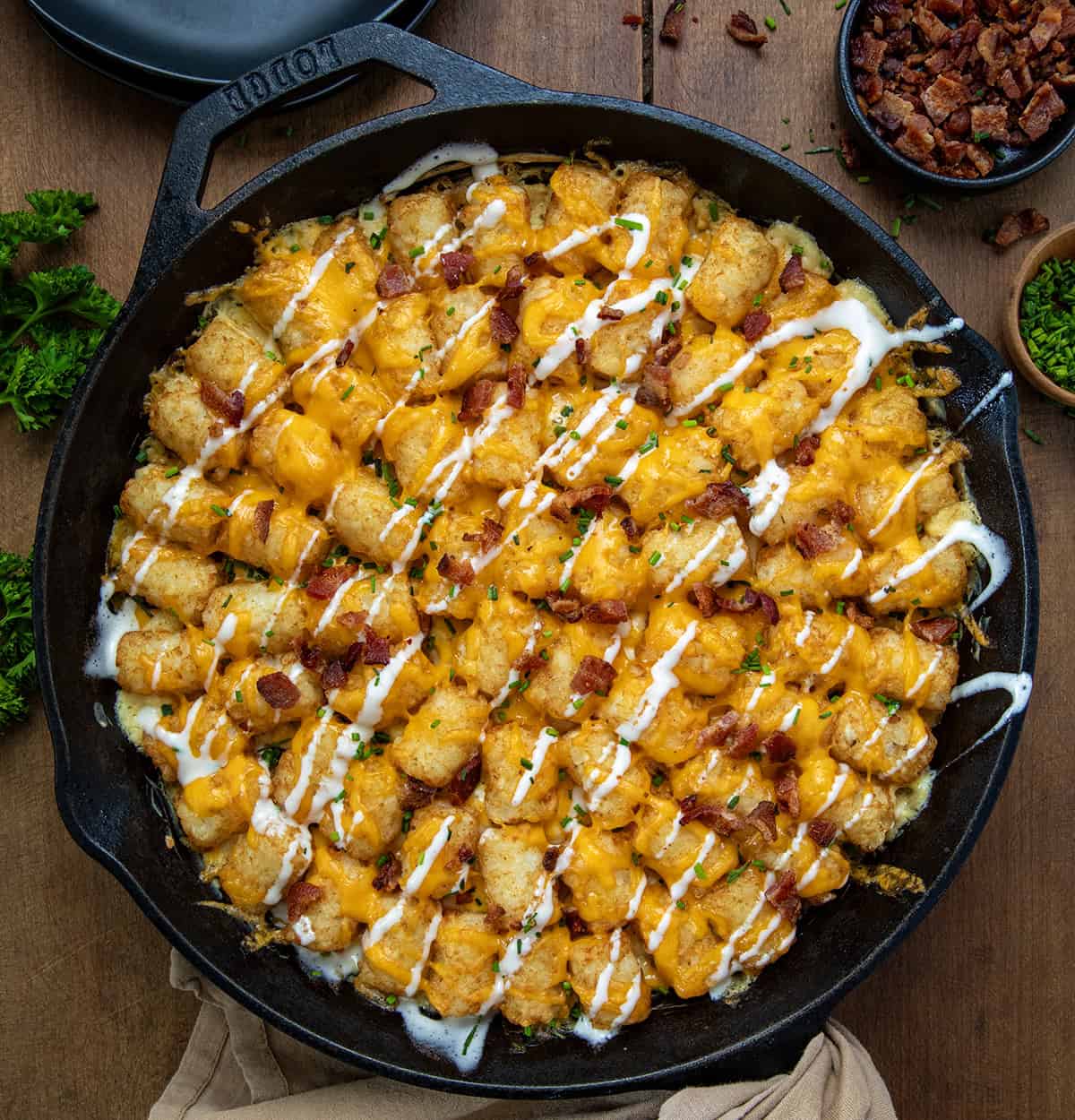 This Chicken Bacon Ranch Tater Tot Casserole is a fun and delicious spin on the classic Tater Tot Hotdish! It replaces ground beef and vegetables with rotisserie chicken, crispy bacon, melty cheddar, and a creamy ranch-seasoned base. Topped with tater tots, more bacon, and a sprinkle of chives, it’s cozy, packed with flavor, and perfect with a drizzle of ranch dressing to tie all of the flavors together! Skillet of Chicken Bacon Ranch Tater Tot Casserole on a wooden table from overhead.