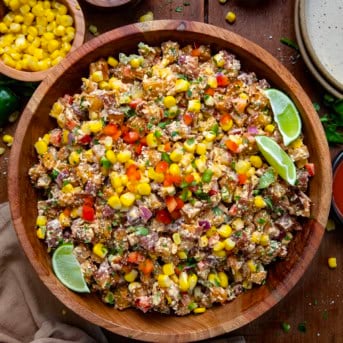 Wooden bowl filled with Street Corn Roasted Potato Salad on a wooden table surrounded by corn and fresh limes.