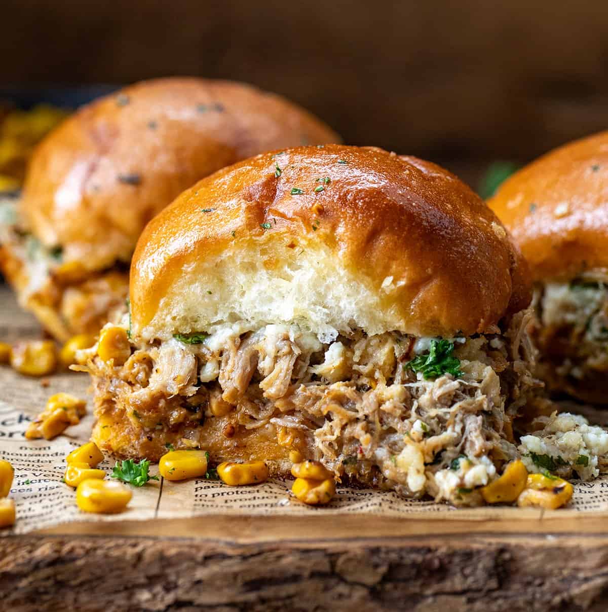 These Carnitas Street Corn Sliders are layered with creamy, smoky street corn, tender shredded carnitas, and plenty of melty cheese, all tucked inside soft dinner rolls and brushed with garlic butter. Baked until golden and gooey, they’re perfect for game day, parties, or a crowd-pleasing weeknight dinner! Close up of a Carnitas Street Corn Slider on a cutting board with more in the background.