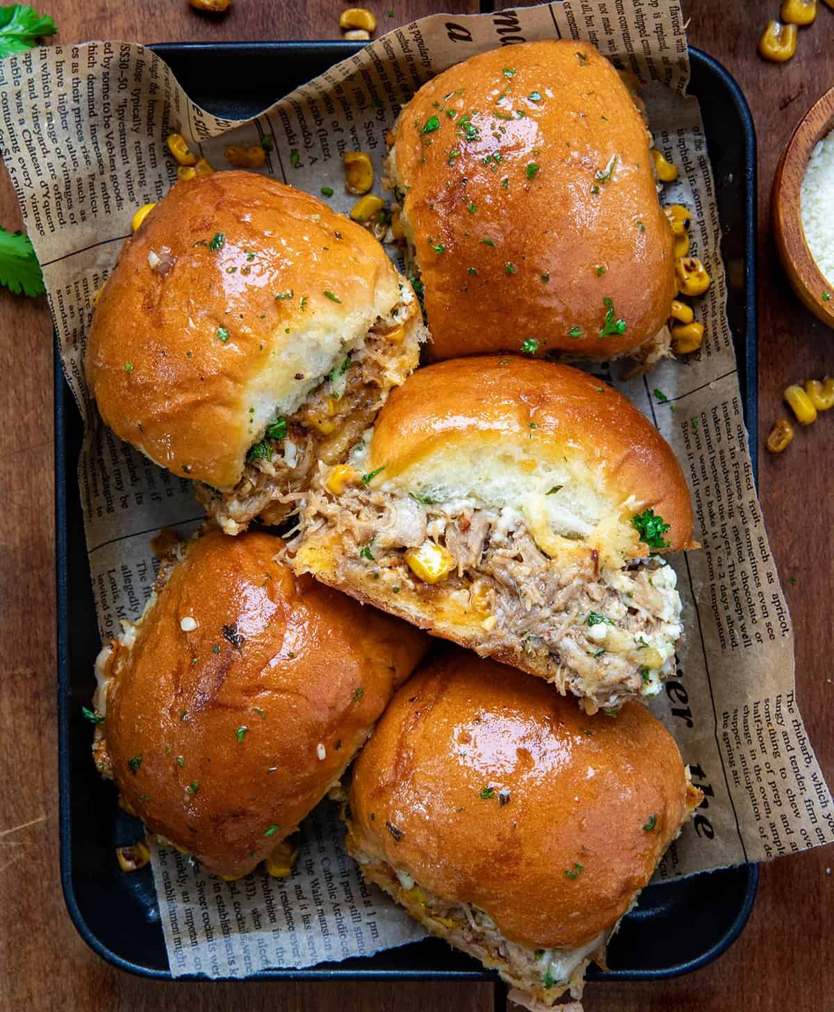 These Carnitas Street Corn Sliders are layered with creamy, smoky street corn, tender shredded carnitas, and plenty of melty cheese, all tucked inside soft dinner rolls and brushed with garlic butter. Baked until golden and gooey, they’re perfect for game day, parties, or a crowd-pleasing weeknight dinner! Carnitas Street Corn Sliders in a small black platter on a wooden table from overhead.