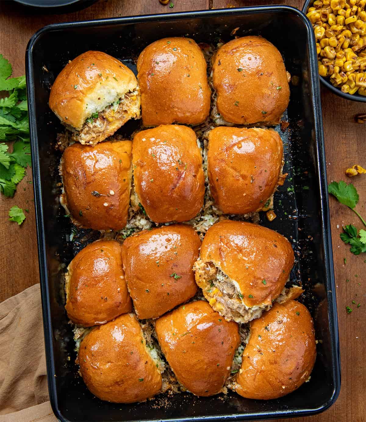 These Carnitas Street Corn Sliders are layered with creamy, smoky street corn, tender shredded carnitas, and plenty of melty cheese, all tucked inside soft dinner rolls and brushed with garlic butter. Baked until golden and gooey, they’re perfect for game day, parties, or a crowd-pleasing weeknight dinner! Black pan of Carnitas Street Corn Sliders on a wooden table from overhead.