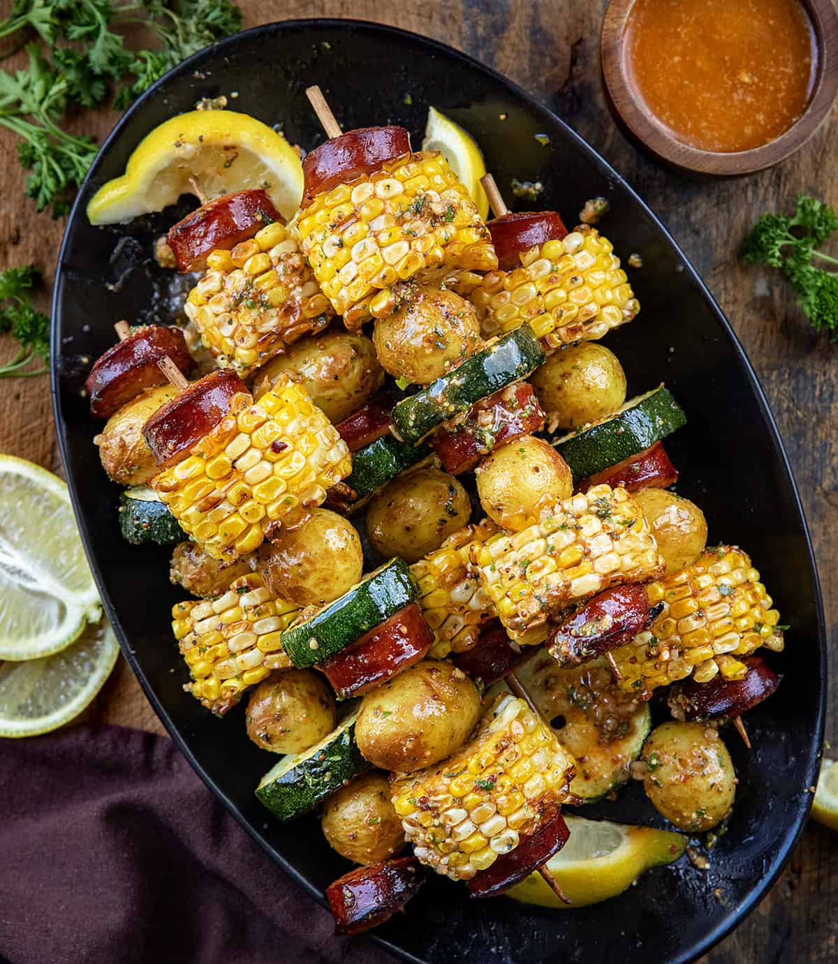 Black platter of Cowboy Butter Kielbasa and Potato Skewers on a wooden table with fresh lemon from overhead.