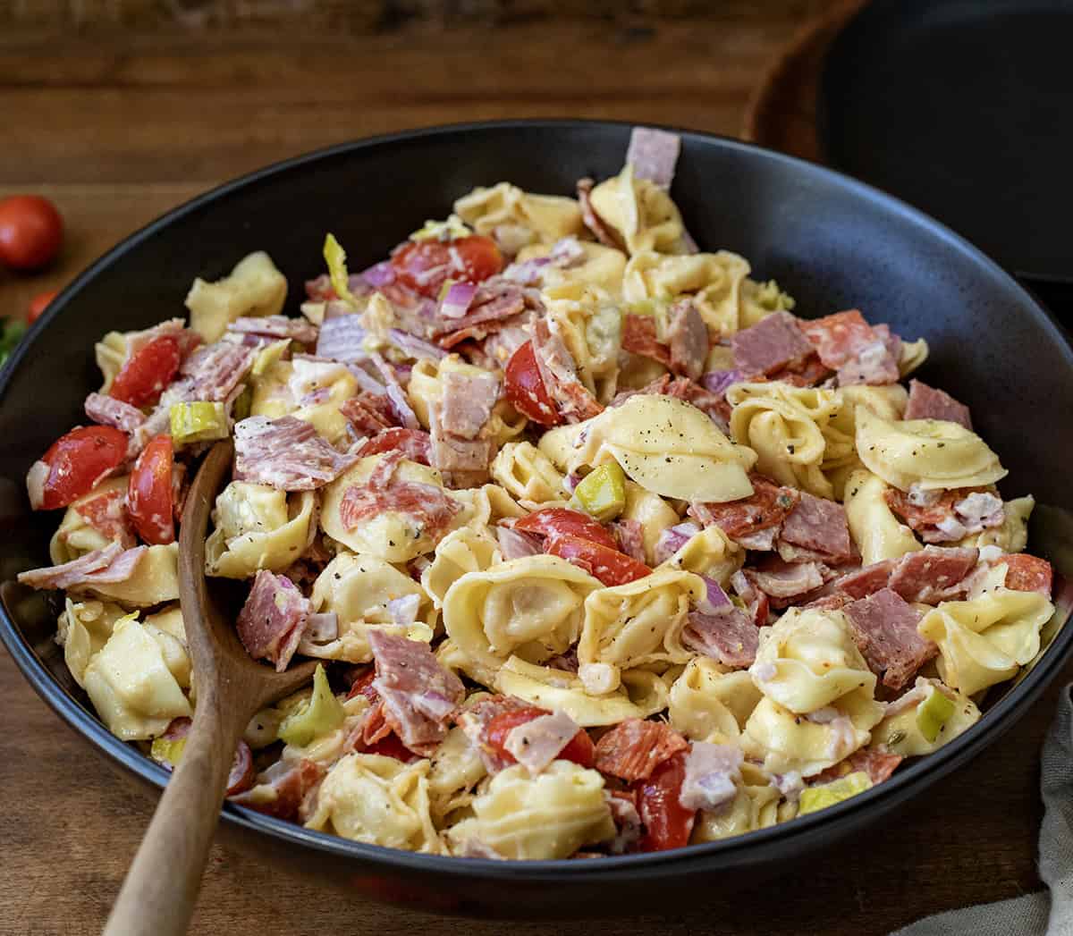 Big black bowl of Italian Grinder Tortellini Salad on a wooden table with a wooden spoon in the bowl.