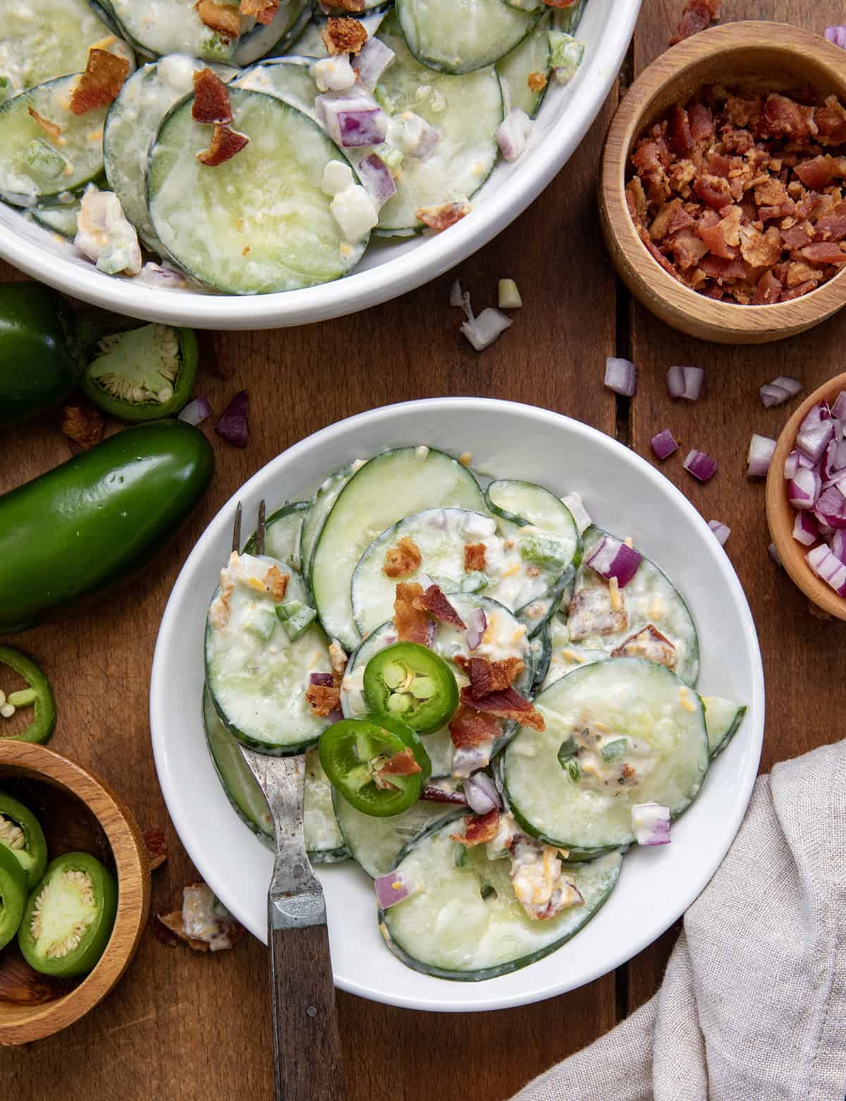 Small white bowl of Jalapeno Popper Cucumber Salad next to a larger bowl on a wooden table.