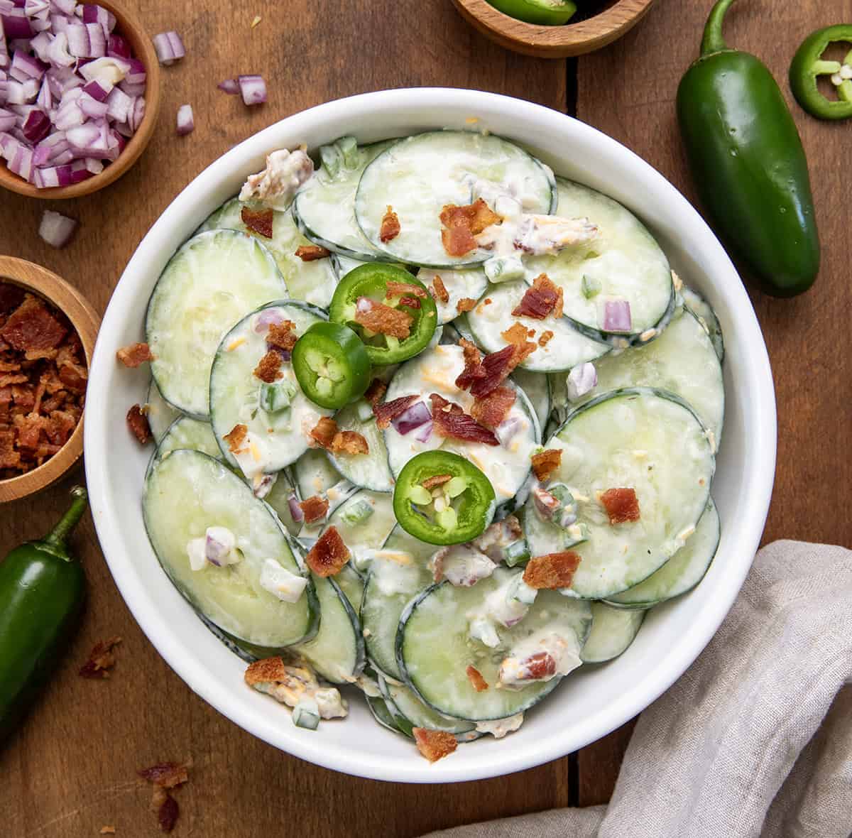 This Jalapeño Popper Cucumber Salad is a cool, creamy, and somewhat spicy side dish perfect for summer cookouts or easy snacks. This easy summer salad is quick to make, super fresh, and tastes even better chilled! Bowl of Jalapeno Popper Cucumber Salad on a wooden table from overhead.
