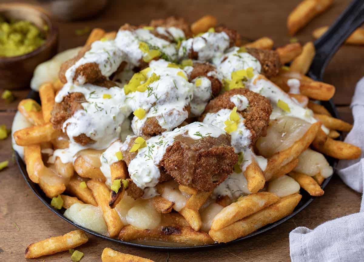This Pickle Poutine is a cheesy, tangy mash-up you didn't know you needed! (Well, I didn't, at least, but now I do!) Crispy fries get smothered in hot brown gravy and melty cheese curds, then topped with crunchy fried pickles and a drizzle of dill pickle ranch! Skillet of Pickle Poutine on a wooden table with french fries and chopped pickles.
