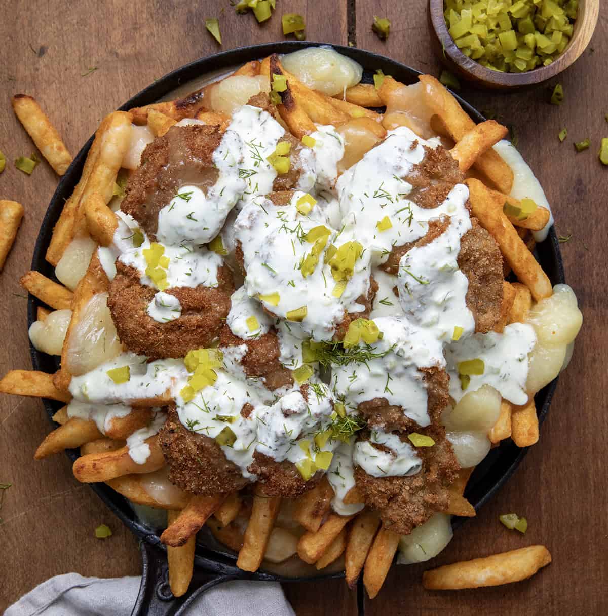This Pickle Poutine is a cheesy, tangy mash-up you didn't know you needed! (Well, I didn't, at least, but now I do!) Crispy fries get smothered in hot brown gravy and melty cheese curds, then topped with crunchy fried pickles and a drizzle of dill pickle ranch! Skillet of Pickle Poutine on a wooden table with french fries and chopped pickles.