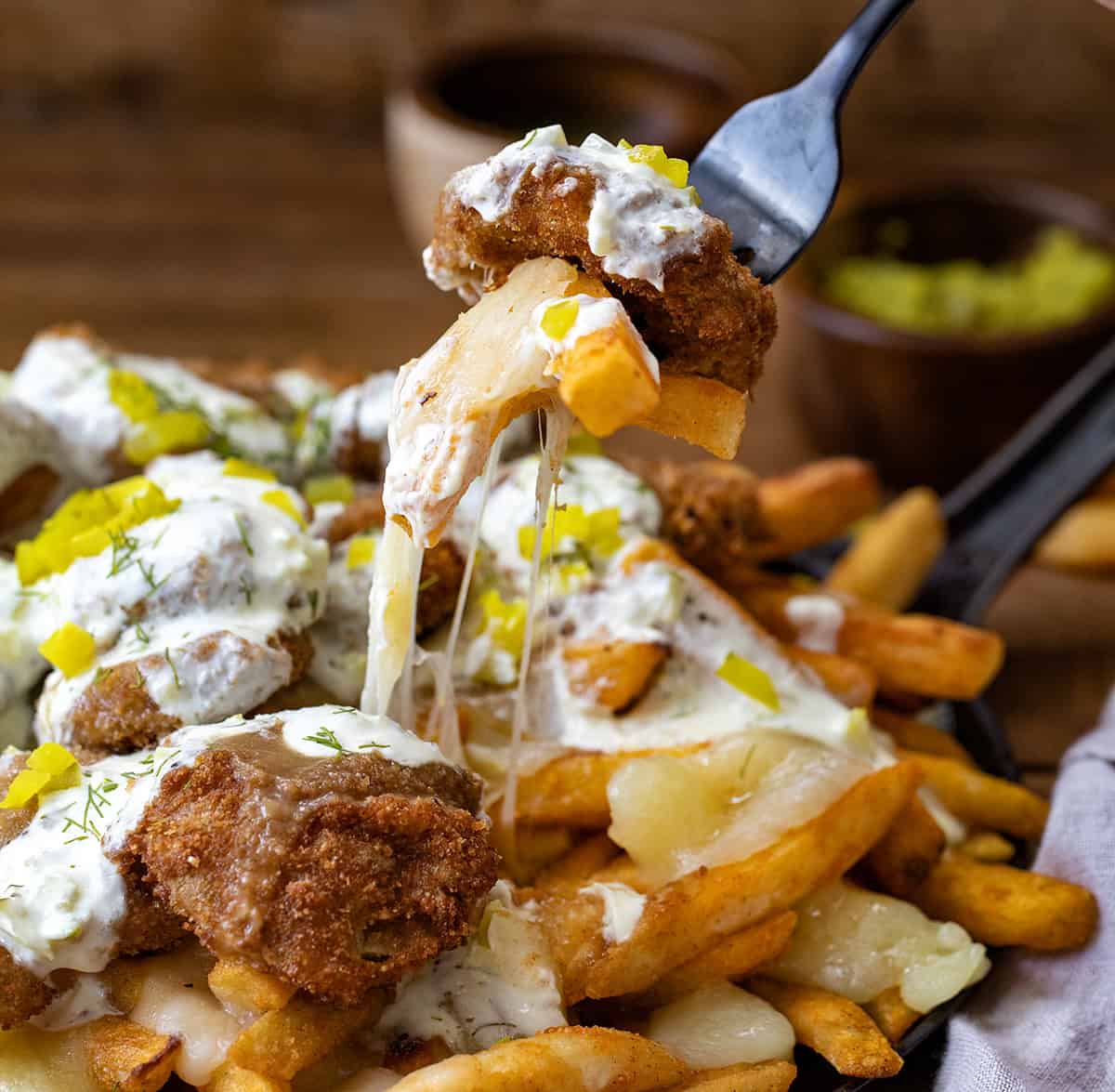 This Pickle Poutine is a cheesy, tangy mash-up you didn't know you needed! (Well, I didn't, at least, but now I do!) Crispy fries get smothered in hot brown gravy and melty cheese curds, then topped with crunchy fried pickles and a drizzle of dill pickle ranch! Fork picking up a cheesy bite of Pickle Poutine.