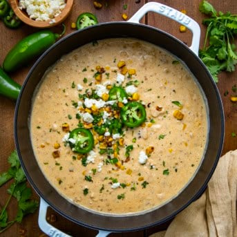 Pot of Street Corn White Chicken Chili on a wooden table surrounded by jalapeno peppers and cheese.