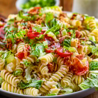 Tossing a BLT Pasta Salad in a white bowl on a wooden table.