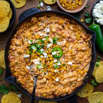 Street Corn Queso in a skillet with a spoon on a wooden table surrounded with chips and roasted corn from overhead.