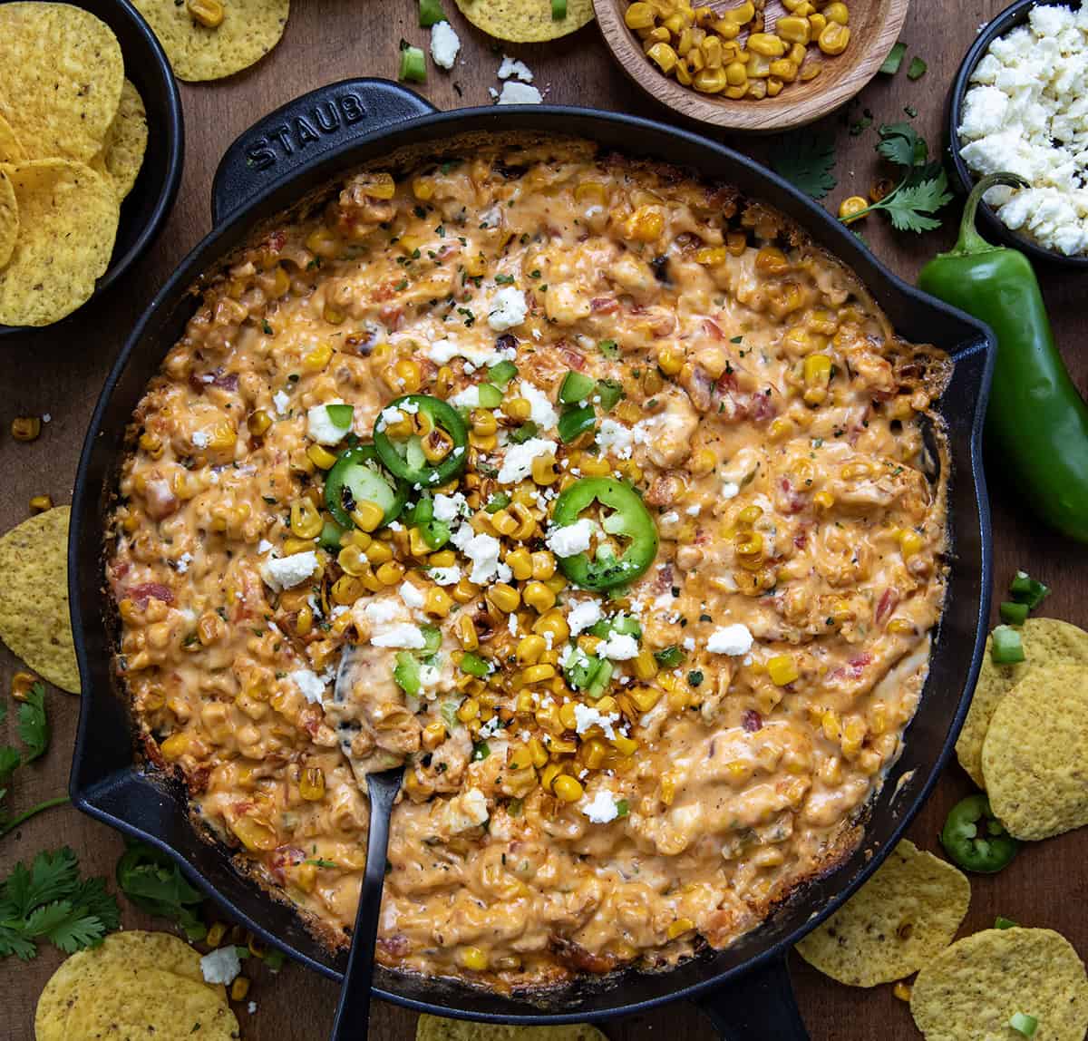 Street Corn Queso in a skillet with a spoon on a wooden table surrounded with chips and roasted corn from overhead.
