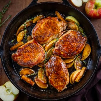 Skillet of Apple Cider Pork Chops on a wooden table with fresh apples from overhead.
