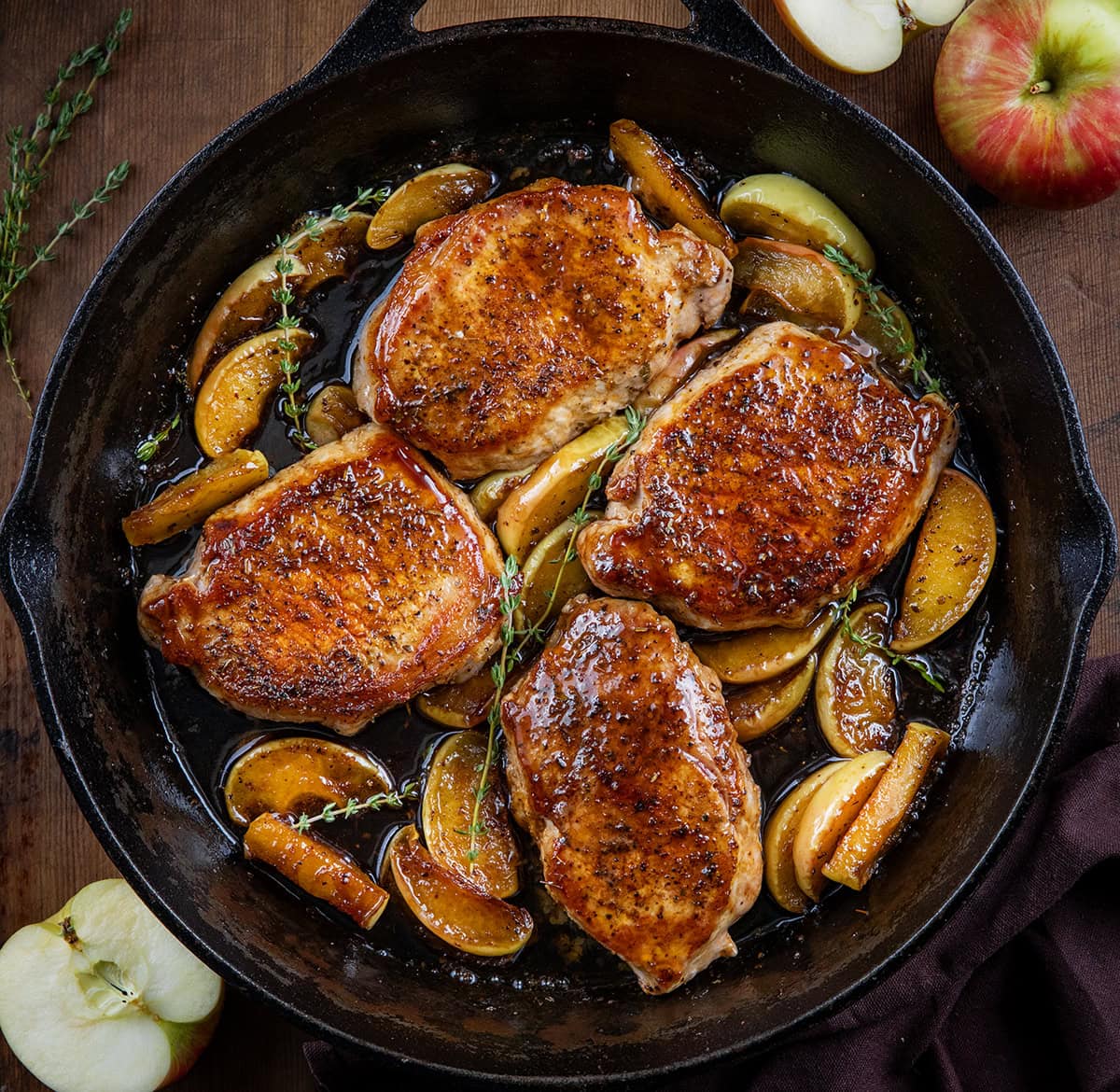 Skillet of Apple Cider Pork Chops on a wooden table with fresh apples from overhead. 