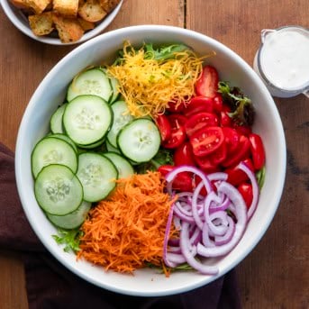 Raw Simple Side Salad ingredients in a bowl next to ranch and homemade croutons on a wooden table from overhead.