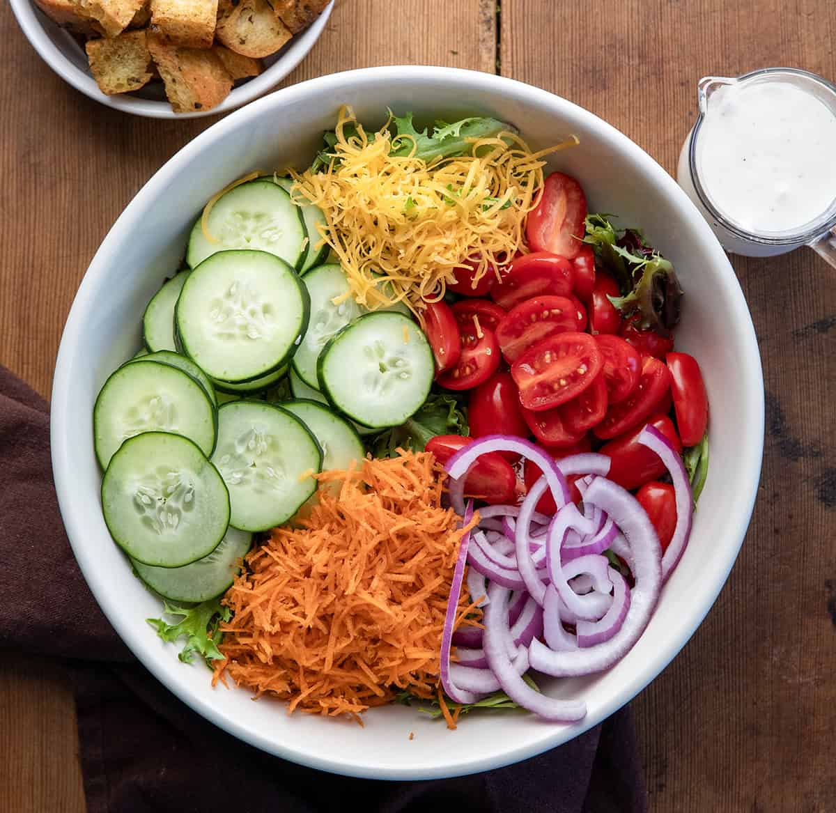 Raw Simple Side Salad ingredients in a bowl next to ranch and homemade croutons on a wooden table from overhead.