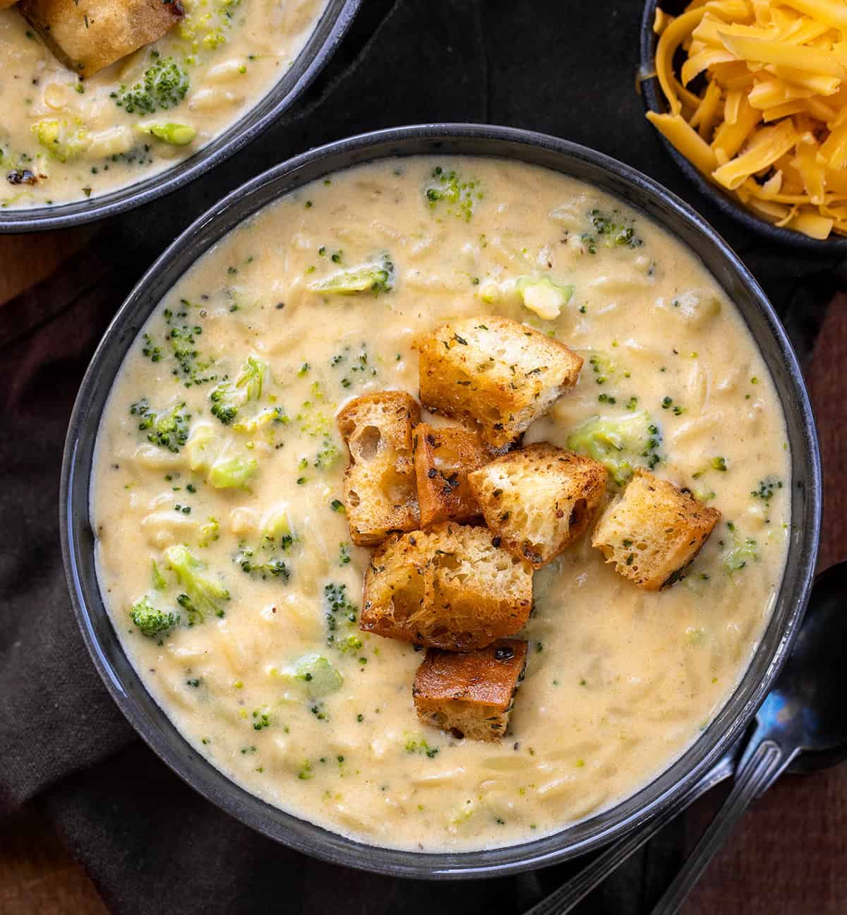 Black bowls of Broccoli Cheddar Orzo Soup with croutons on a wooden table from overhead.
