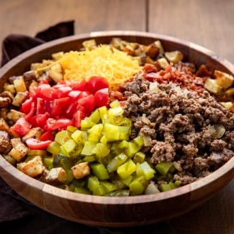 Wooden bowl filled with potatoes, ground beef, pickles, tomatoes, cheese, and bacon to make a Cheeseburger Bowl.