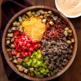 Wooden bowl filled with potatoes, ground beef, pickles, tomatoes, cheese, and bacon to make a Cheeseburger Bowl from overhead.
