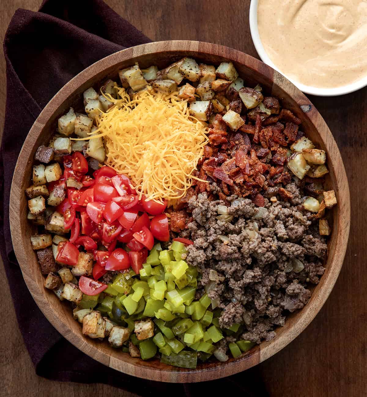 Wooden bowl filled with potatoes, ground beef, pickles, tomatoes, cheese, and bacon to make a Cheeseburger Bowl from overhead.