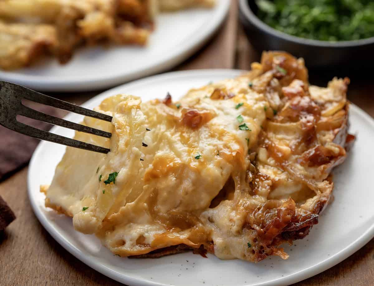 These Cheesy French Onion Scalloped Potatoes are rich, creamy, and loaded with caramelized onions and melty cheese! Fork picking up a portion of cheesy French Onion Scalloped Potatoes from white plate.