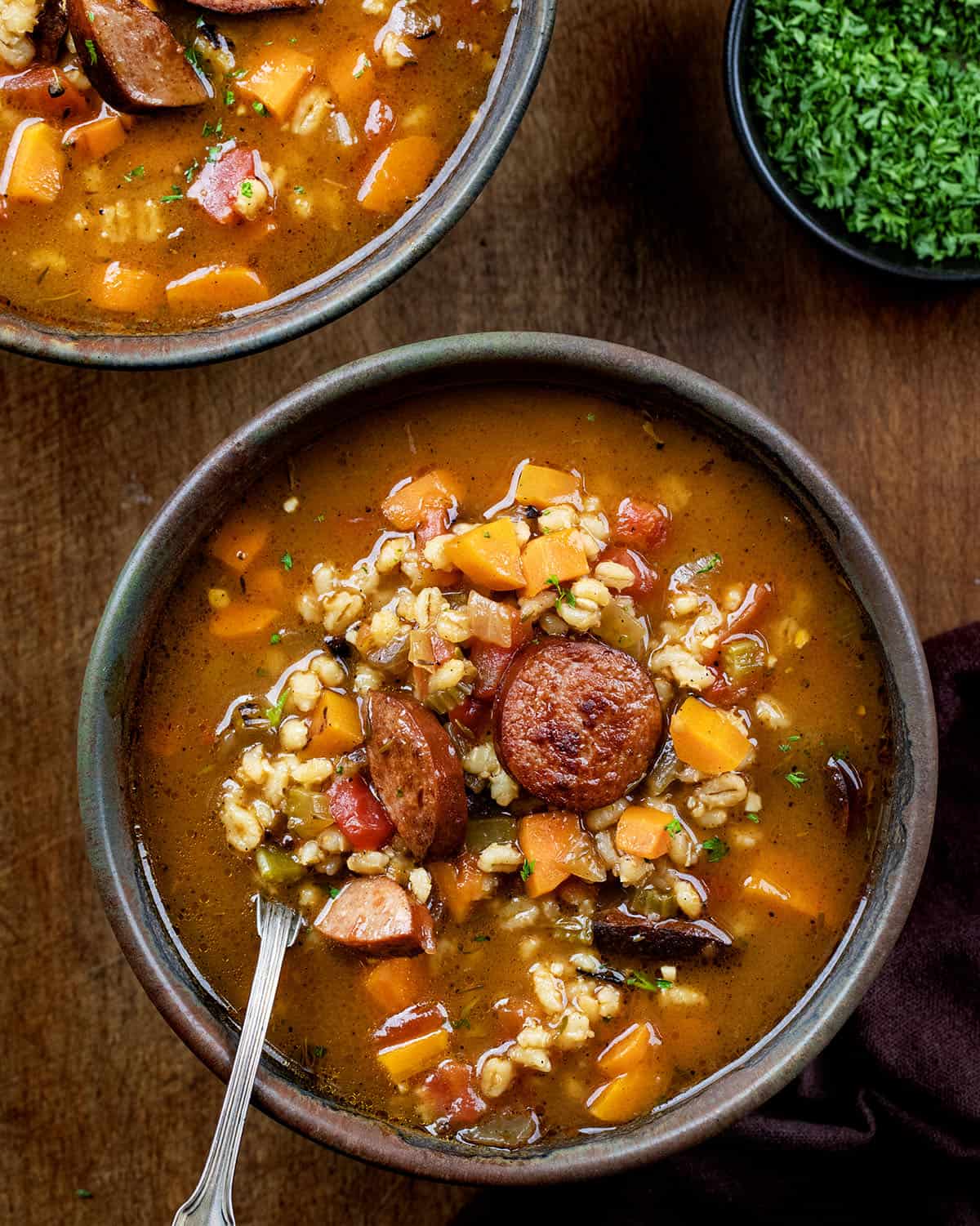 Bowls of Kielbasa Barley Soup one with a spoon in it on a wooden table from overhead.