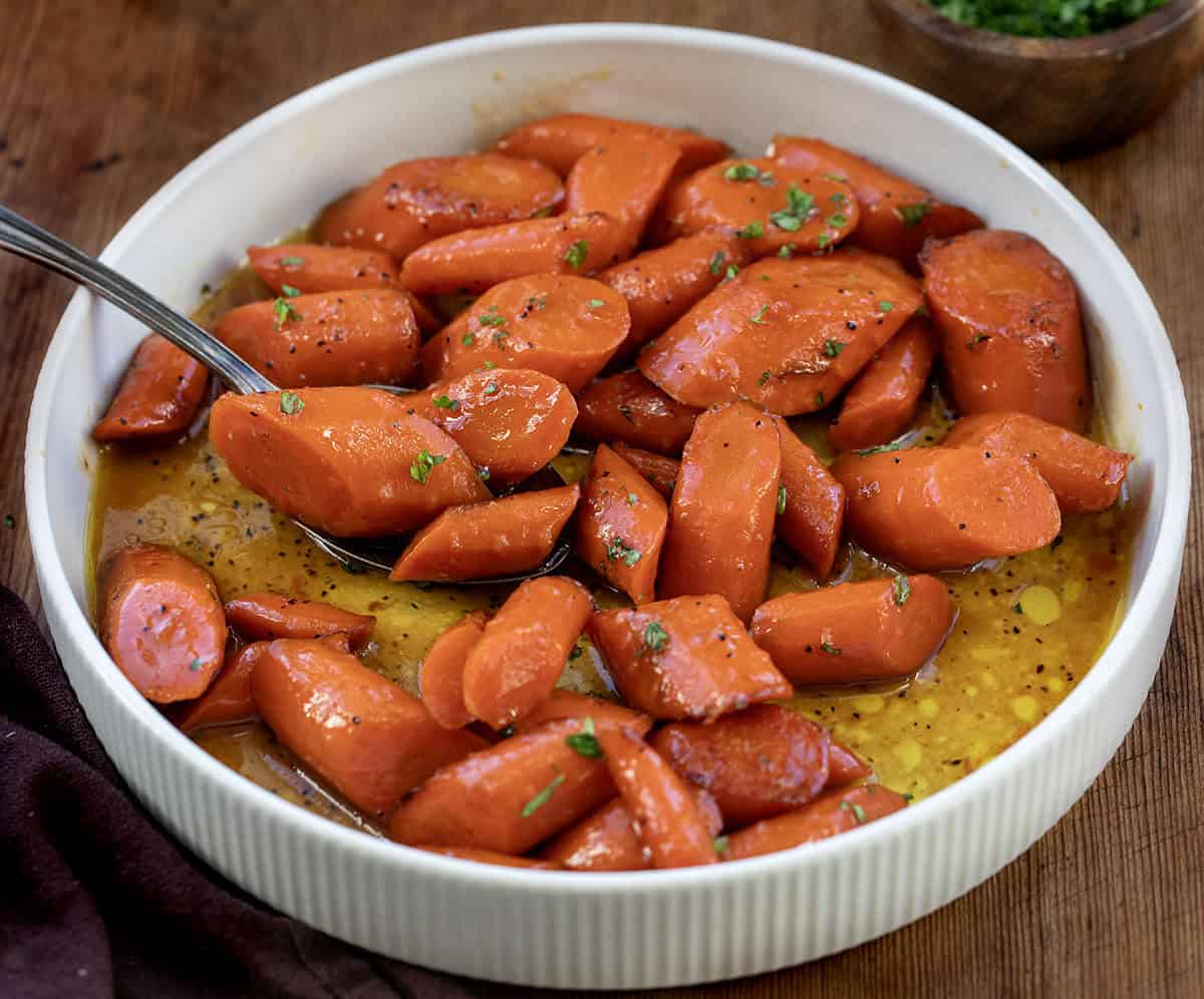 Elevate your holiday table or any weeknight dinner with Candied Carrots, sweet and buttery glazed carrots perfect for any occasion! Cream bowl of Candied Carrots with a spoon.