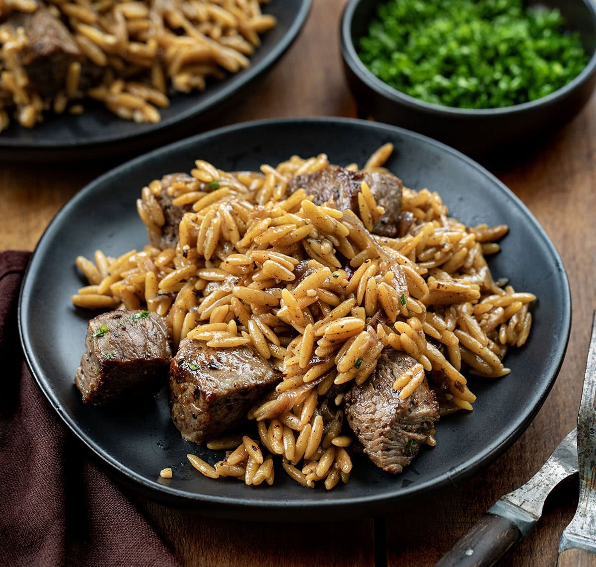 Steak and Caramelized Onion Orzo gives you the best of both worlds. Tender seared steak bites meet deeply sweet caramelized onions, all nestled in creamy Parmesan orzo that feels special enough for company but easy enough for any weeknight! Black plates with Steak and Caramelized Onion Orzo on them.