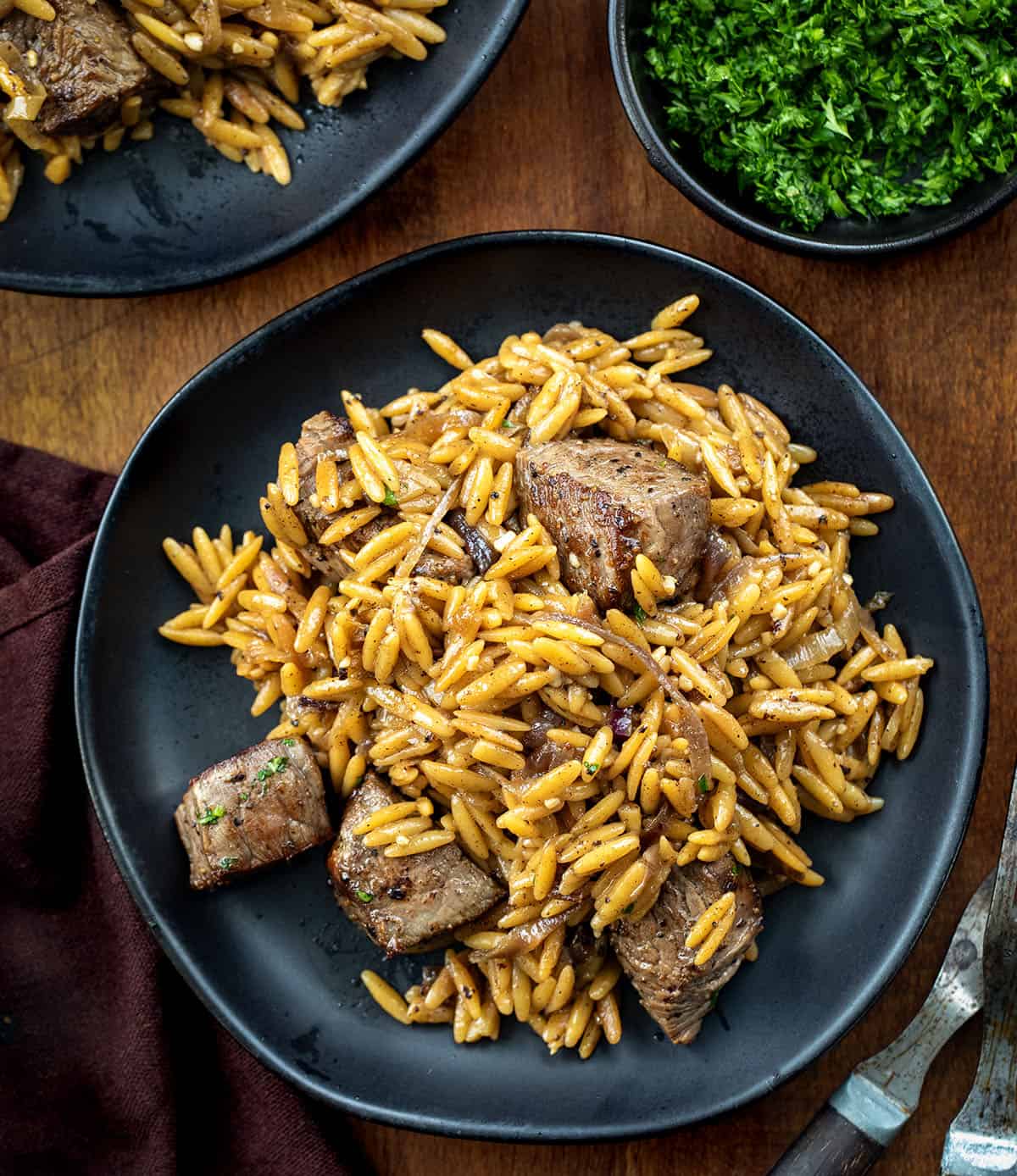 Steak and Caramelized Onion Orzo gives you the best of both worlds. Tender seared steak bites meet deeply sweet caramelized onions, all nestled in creamy Parmesan orzo that feels special enough for company but easy enough for any weeknight! Black plates with Steak and Caramelized Onion Orzo on a wooden table from overhead.