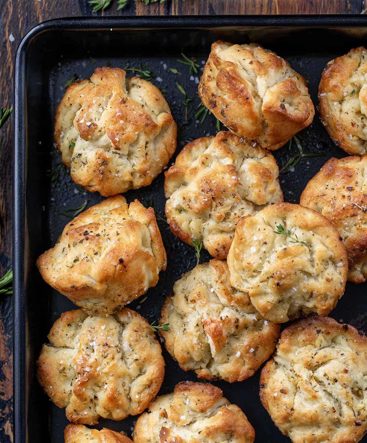 These Focaccia Muffins are crispy on the outside, soft and airy inside, and bursting with garlic, olive oil, and herbs. Focaccia Muffins in a black pan on a wooden table from overhead.