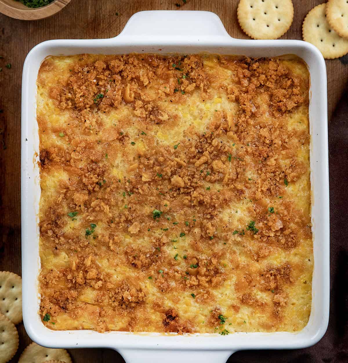 Cozy, creamy, and crowned with a buttery cracker topping, this Scalloped Corn is the kind of classic holiday side that always disappears fast! Scalloped Corn in a white baking dish on a wooden table with Ritz crackers.