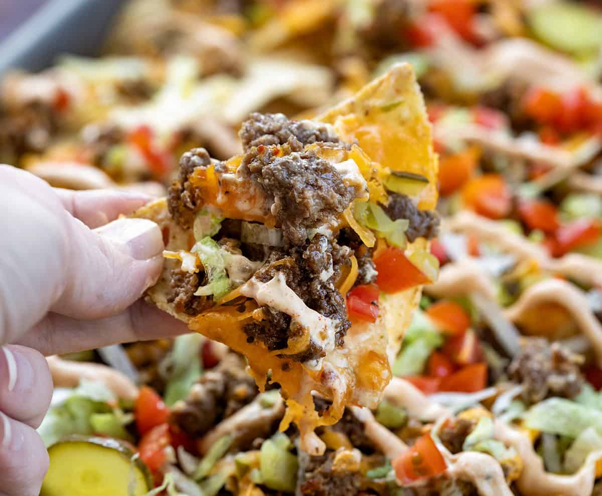 Cheeseburger Nachos are an easy, sheet-pan appetizer that’s perfect for game day or any casual gathering! Hand holding a chip covered in burger, cheese, pickles, and tomatoes.