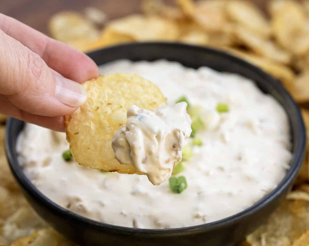 Whip up an irresistible Clam Dip in under 10 minutes! Perfect for gatherings or as a special treat for yourself. Hand holding a chip with Clam Dip on it.