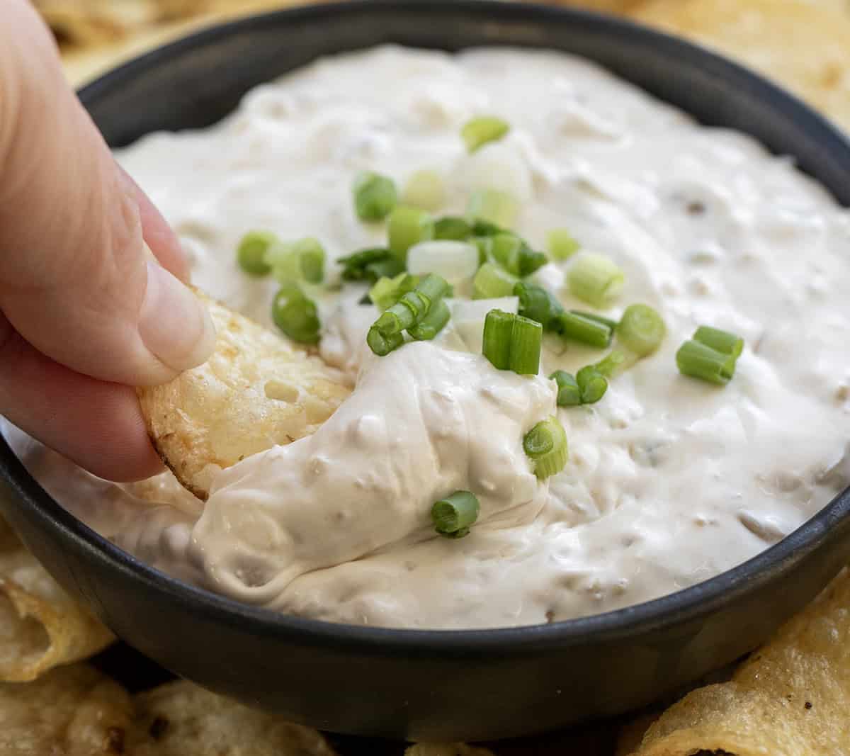 Whip up an irresistible Clam Dip in under 10 minutes! Perfect for gatherings or as a special treat for yourself. Hand holding a chip with Clam Dip on it being scooped out of the bowl.
