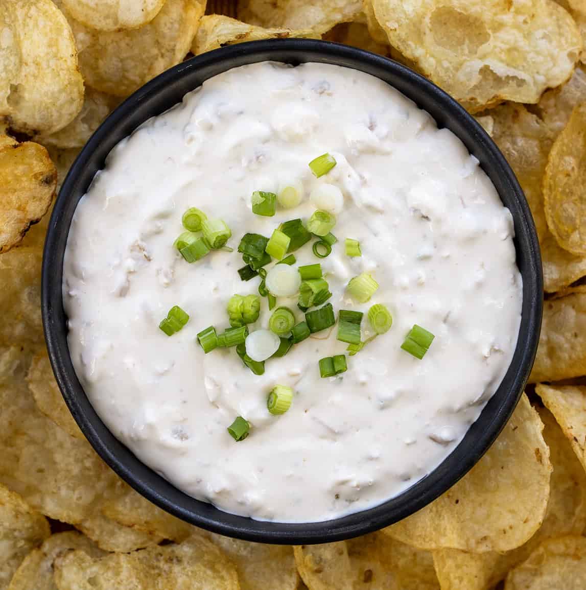Whip up an irresistible Clam Dip in under 10 minutes! Perfect for gatherings or as a special treat for yourself. Small black bowl filled with Clam Dip surrounded by potato chips.