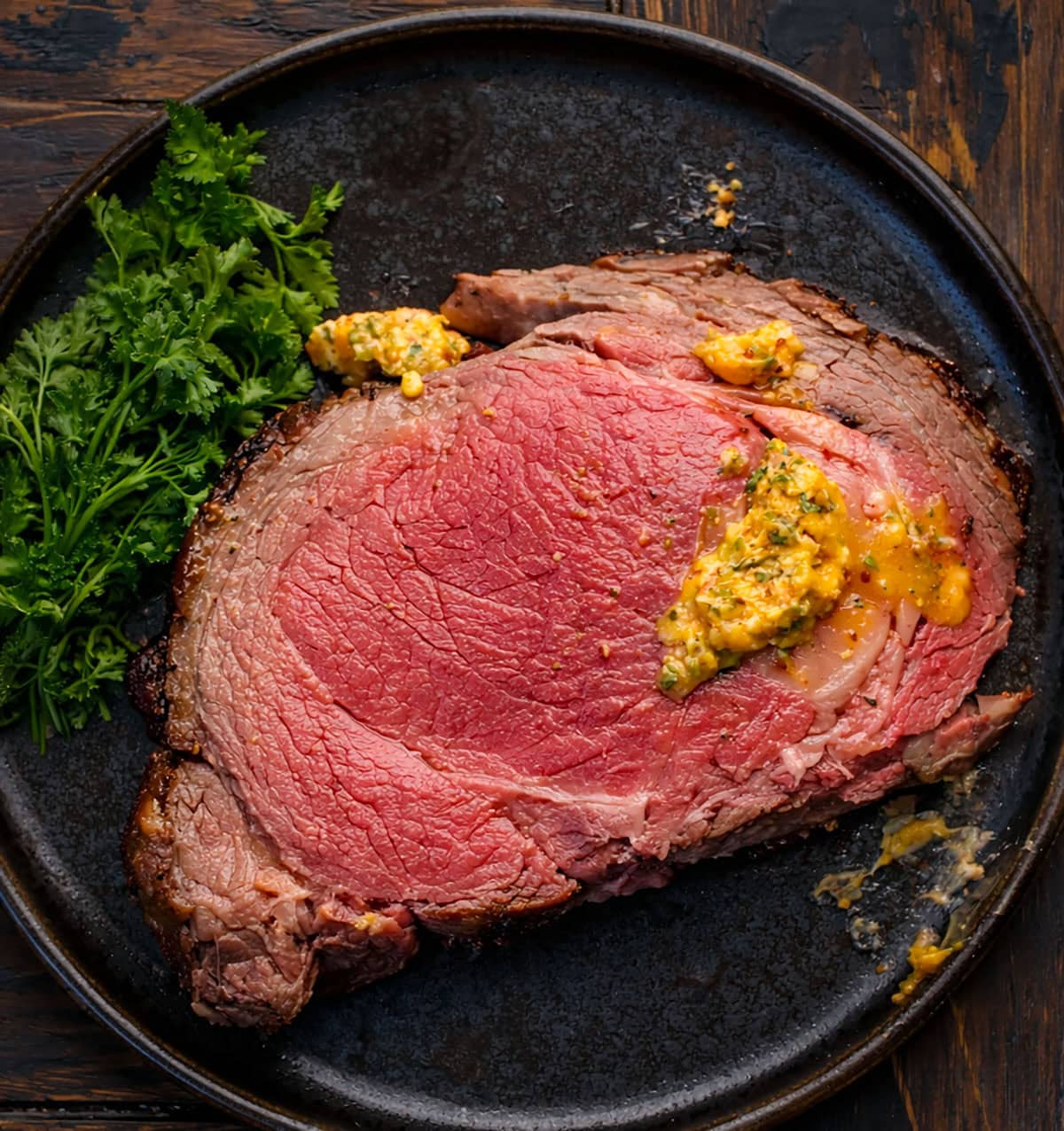 All the flavor of my popular Prime Rib gets an extra punch with my homemade Cowboy Butter! This prime rib is perfectly roasted, with a golden, flavorful crust, and each juicy slice is topped with a spicy, herby butter that melts into the meat! Piece of Cowboy Butter Prime Rib on a black plate on a wooden table from overhhead with some cowboy butter on the meat.