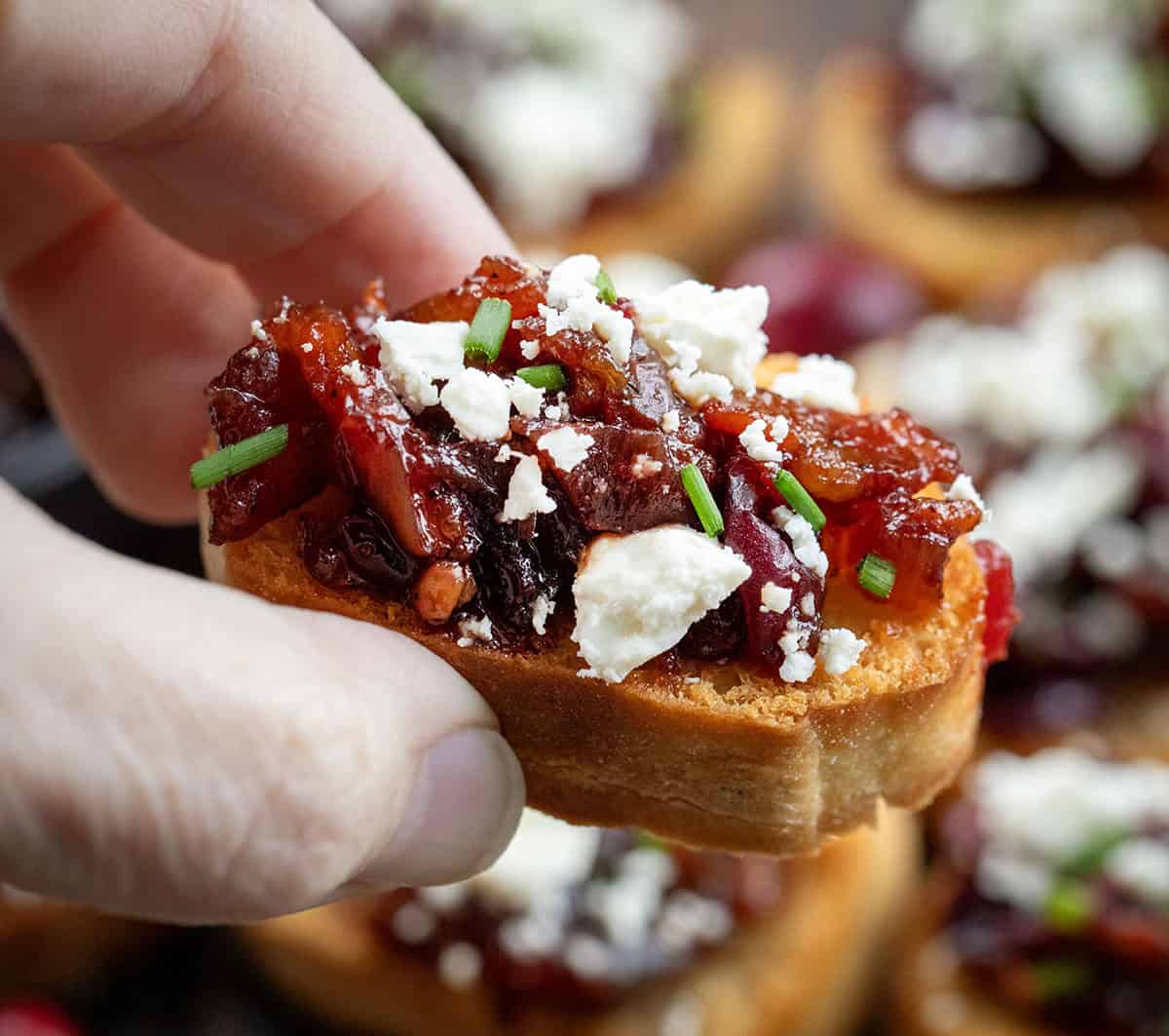 These Cranberry Bacon Jam and Feta Crostini are the ultimate sweet-and-savory appetizer! Crisp, golden baguette slices are topped with my homemade cranberry bacon jam and a sprinkle of creamy feta! Hand holding a Cranberry Bacon Jam and Feta Crostini.