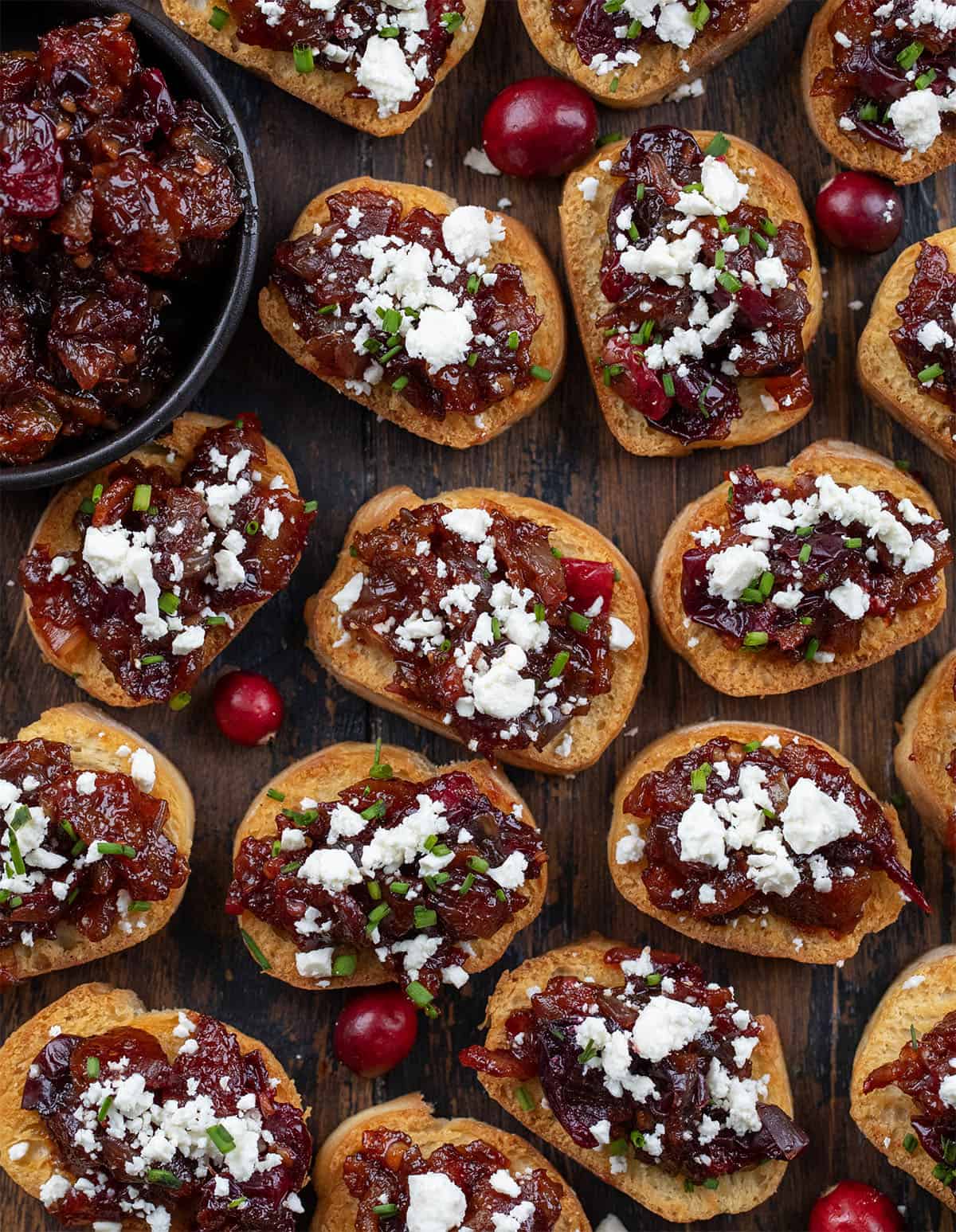 These Cranberry Bacon Jam and Feta Crostini are the ultimate sweet-and-savory appetizer! Crisp, golden baguette slices are topped with my homemade cranberry bacon jam and a sprinkle of creamy feta! Cranberry Bacon Jam and Feta Crostini laid out on a wooden table from overhead.