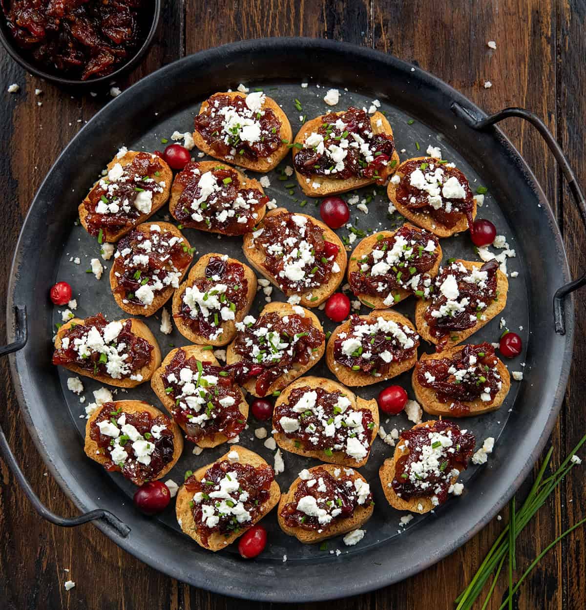 These Cranberry Bacon Jam and Feta Crostini are the ultimate sweet-and-savory appetizer! Crisp, golden baguette slices are topped with my homemade cranberry bacon jam and a sprinkle of creamy feta! Platter of Cranberry Bacon Jam and Feta Crostini on a wooden table from overhead.