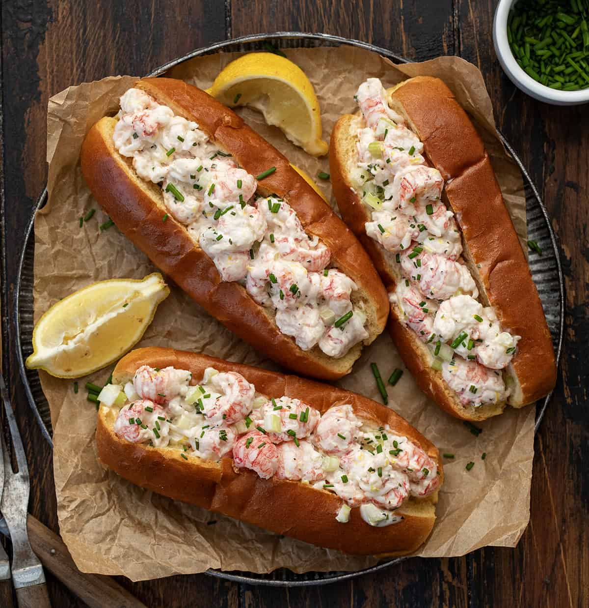 These Langostino Lobster Rolls are my quick, easy take on a classic New England favorite. Sweet, tender langostino takes the place of lobster tails, tossed in a bright, lemony mayo with celery and chives, then served in buttery toasted brioche buns! Platter of Langostino Lobster Rolls with lemon slices on a wooden table from overhead.