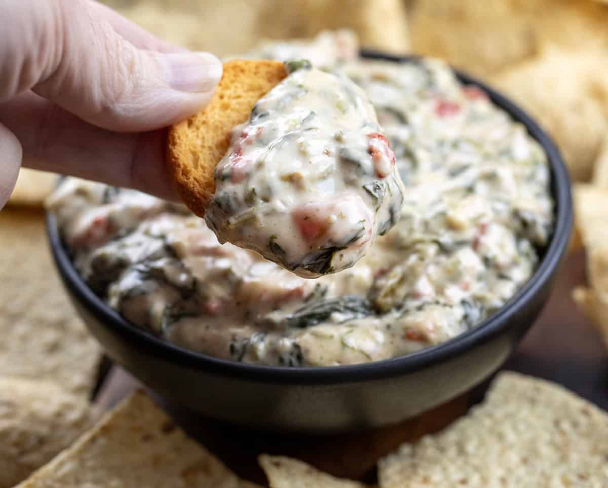Small black bowl of Spinach Queso Dip surrounded by tortilla chips and with a hand holding a dip covered chip above it.