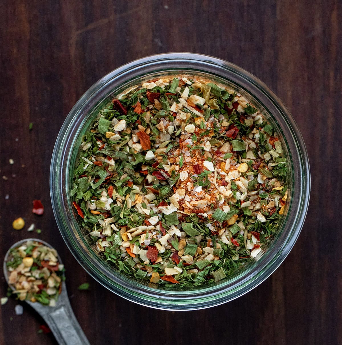 This Cowboy Butter Seasoning is a bold, savory, and slightly spicy blend of herbs and spices that transforms butter, mayonnaise, or cream cheese into flavorful spreads! Jar of Cowboy Butter Seasoning on a wooden table with a metal teaspoon next to the jar from overhead.