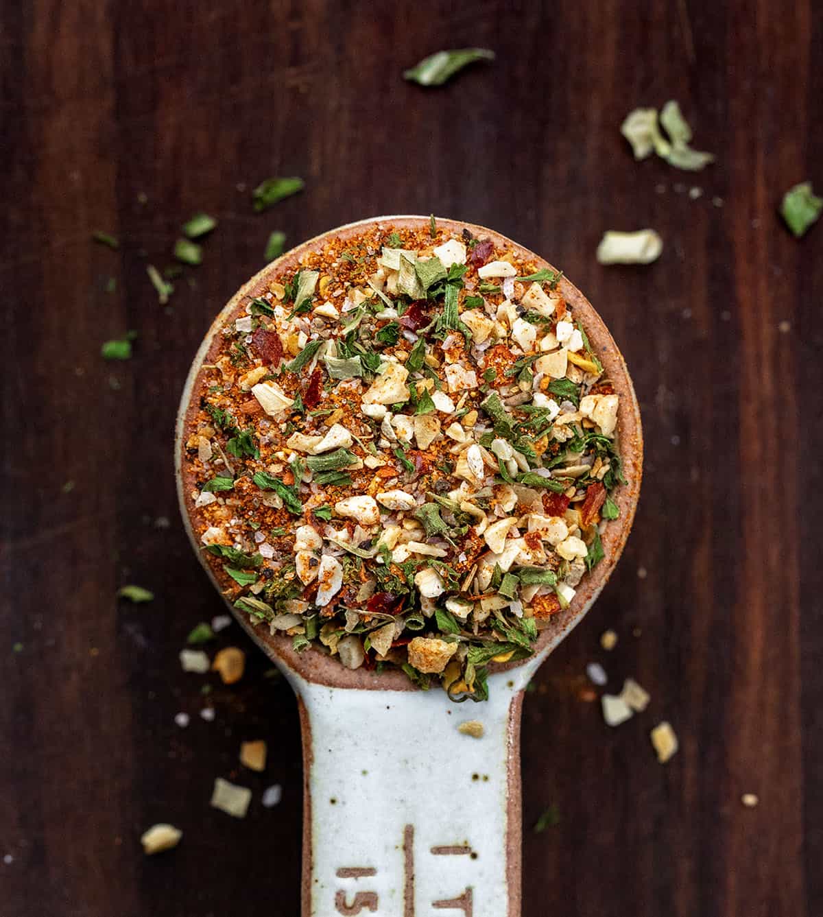 This Cowboy Butter Seasoning is a bold, savory, and slightly spicy blend of herbs and spices that transforms butter, mayonnaise, or cream cheese into flavorful spreads! Ceramic tablespoon filled with Cowboy Butter Seasoning on a wooden table.