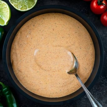 Bowl of Southwest Ranch Sauce with a spoon in it and fresh limes, tomatoes, and jalapeno next to it.