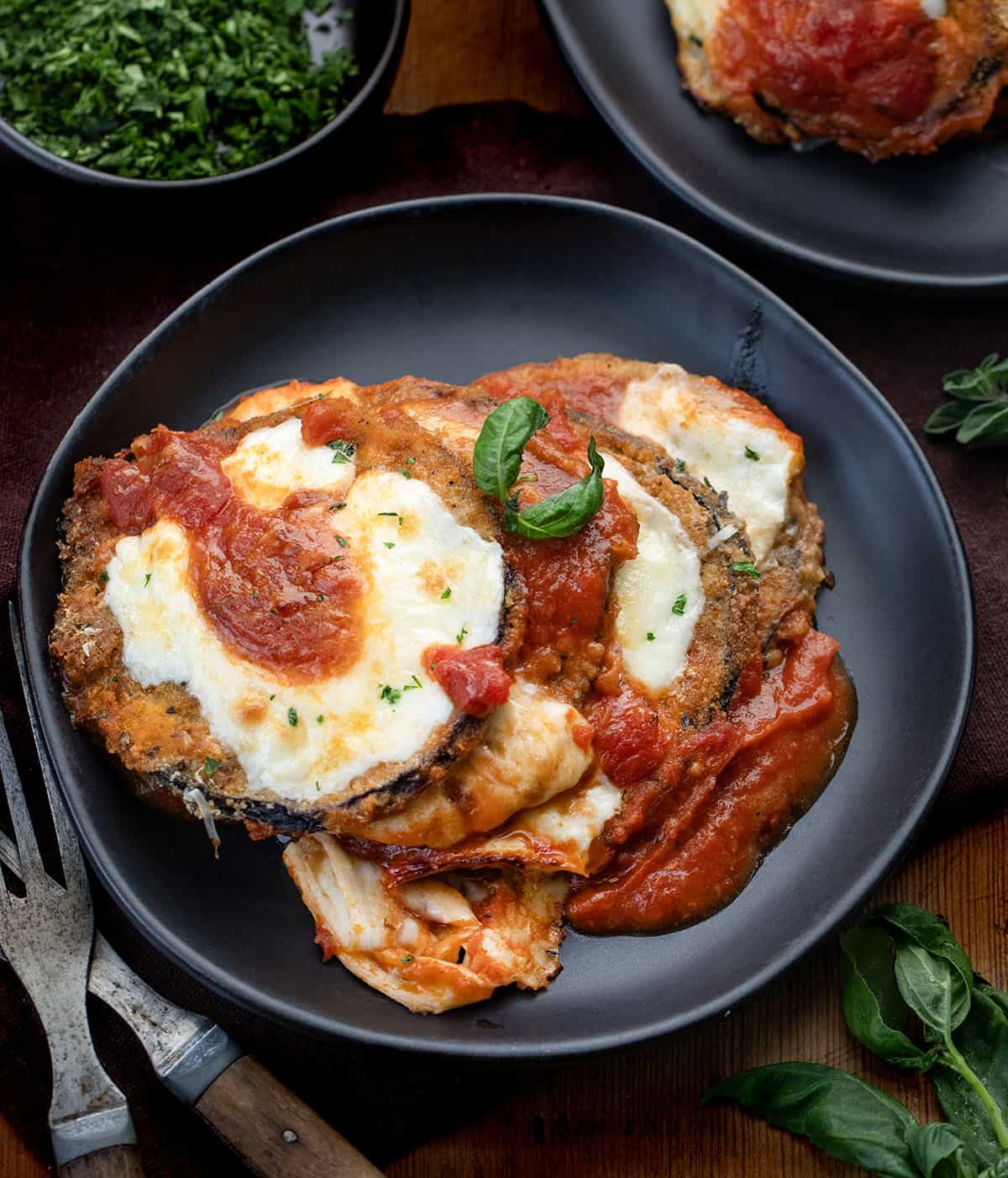 Crispy, golden eggplant slices are layered with rich marinara, melty mozzarella, and savory Parmesan, then baked until bubbly and perfectly browned. This classic Eggplant Parmesan delivers incredible texture in every bite and makes a comforting, crowd-pleasing dinner any night of the week. Eggplant Parmesan on a black plate on a wooden table from overhead.
