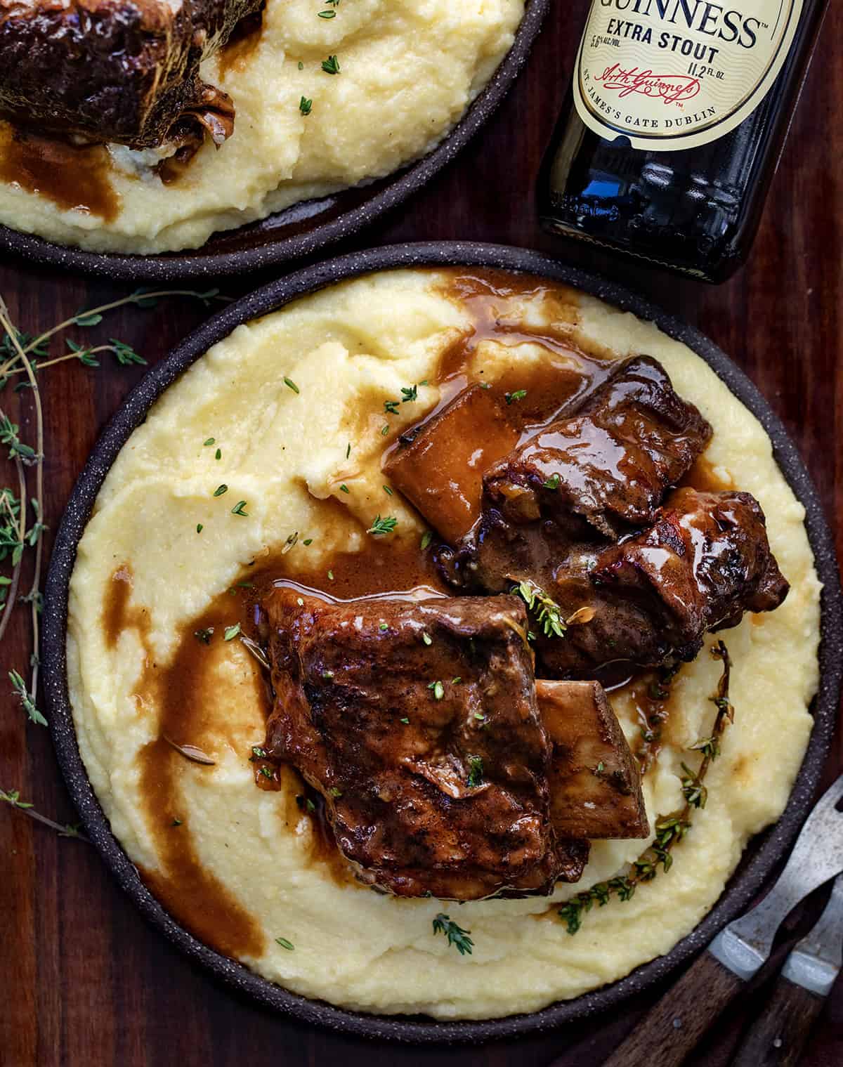 Rich, savory, and slow-braised to perfection, these ribs are cooked in bold Guinness stout, fresh herbs, and a luscious beef broth. The silky pan sauce is perfect spooned over mashed potatoes or noodles, all in one pot for easy, impressive comfort food. Plates of Guinness Short Ribs on polenta on a wooden table.