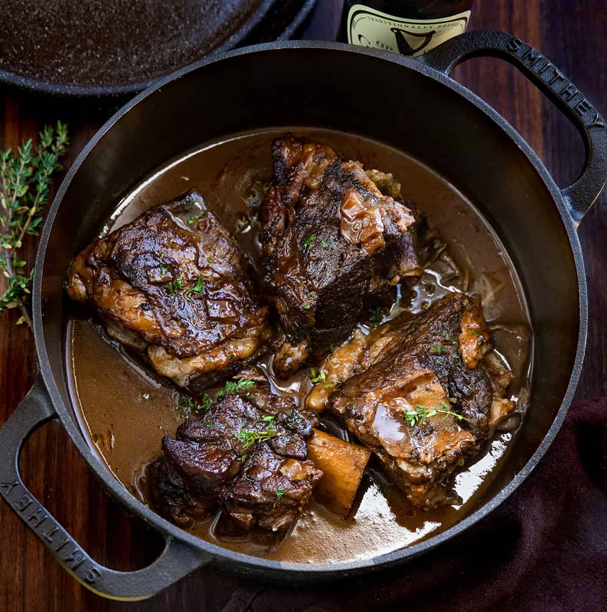 Rich, savory, and slow-braised to perfection, these ribs are cooked in bold Guinness stout, fresh herbs, and a luscious beef broth. The silky pan sauce is perfect spooned over mashed potatoes or noodles, all in one pot for easy, impressive comfort food. Pot of Guinness Short Ribs on a wooden table from overhead.