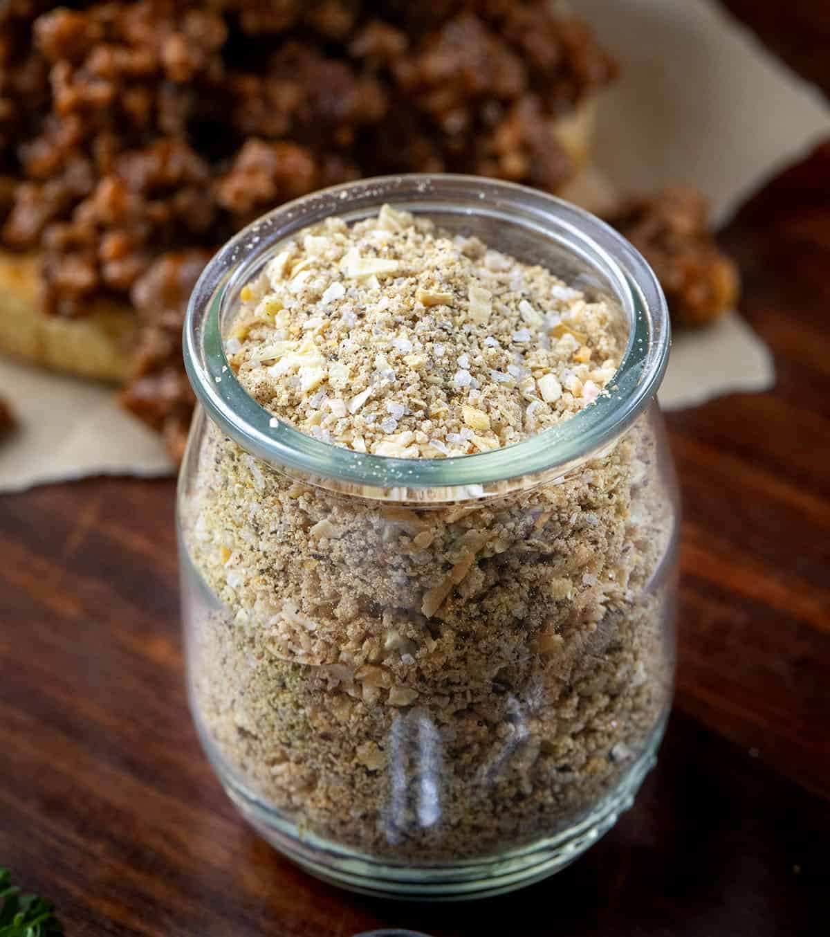 Homemade Sloppy Joe Dry Mix is a simple, pantry-friendly mix that you can whip up in minutes and keep on hand for months. Just a few tablespoons of this mix and ground beef turns into rich, savory, slightly sweet Sloppy Joes that everyone will love! Jar of Homemade Sloppy Joe Dry Mix on a wooden table in front of a sloppy joe sandwich.