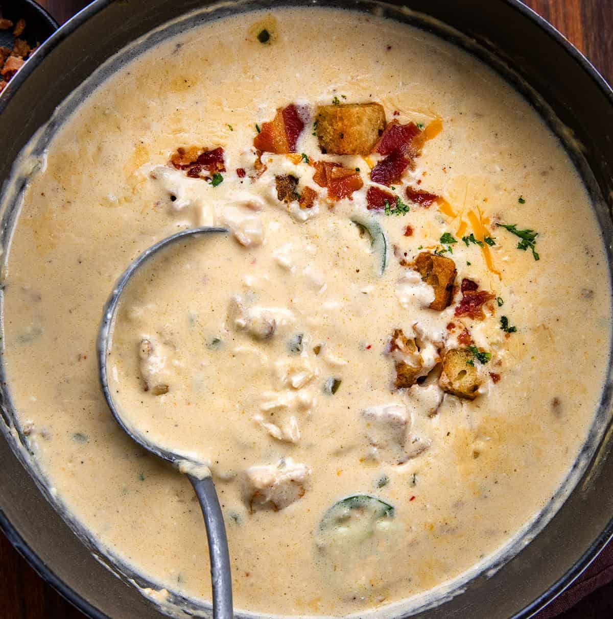 Creamy, cheesy, and just the right amount of spice! This Jalapeño Popper Chicken Soup is packed with tender chicken, crispy bacon, gooey cheese, and a kick of jalapeño. Perfect for cozy nights, weeknight dinners, or anytime you want a comforting, hearty bowl of deliciousness. Pot full of Jalapeño Popper Chicken Soup with a ladle in the pot on a wooden table.