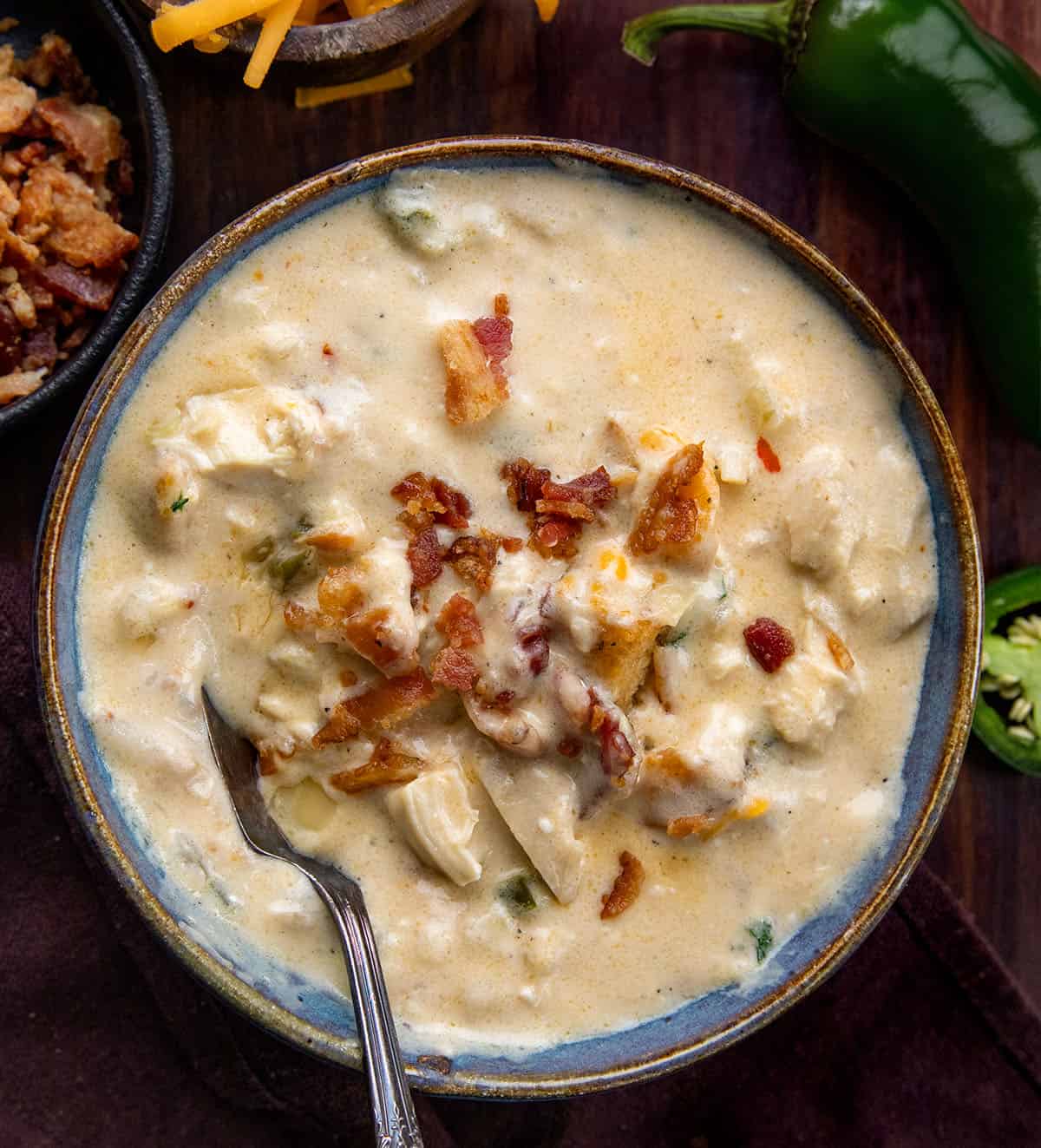 Creamy, cheesy, and just the right amount of spice! This Jalapeño Popper Chicken Soup is packed with tender chicken, crispy bacon, gooey cheese, and a kick of jalapeño. Perfect for cozy nights, weeknight dinners, or anytime you want a comforting, hearty bowl of deliciousness. Bowl of Jalapeño Popper Chicken Soup with a spoon in it on a wooden table from overhead.